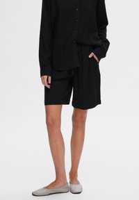 Chemise en lin noire avec fermeture à bouton, associée à un short noir avec taille élastique et poches latérales, portée avec des chaussures slip-ons gris clair.