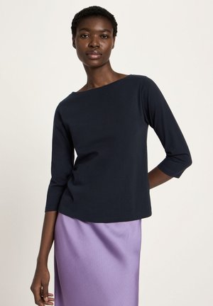 Frau, die ein marineblaues Dreiviertelarmshirt und einen hellvioletten Rock trägt, vor einem schlichten hellen Hintergrund stehend.