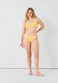 Geel bikini met patroon en witte accenten; triangel top en low-rise broekje, gemaakt van gladde stof, met verstelbare koordjes aan de zijkanten.