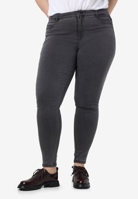 Persona con jeans ajustados gris oscuro y zapatos de cordones rojo oscuro, de pie contra un fondo blanco liso.