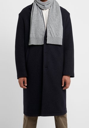 Homme portant un pardessus bleu marine foncé, un pantalon beige, une chemise blanche et une écharpe gris clair drapée autour du cou, sur un fond uni.