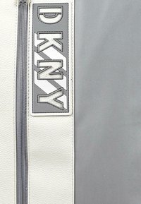 Logo de DKNY en un parche rectangular con una combinación de colores en gris y blanco. Presenta una superficie texturizada y un detalle de cremallera central.