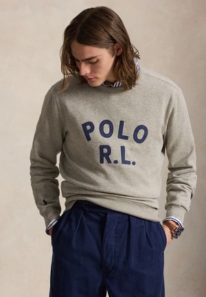 Młody mężczyzna z długimi włosami, ubrany w szarą bluzę z napisem "POLO R.L.", niebieskie spodnie i zegarek na nadgarstku, ręce w kieszeniach, patrzy w dół.