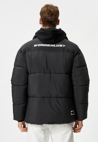 Zwarte pufferjack met een glanzende afwerking, voorzien van een "WONDERLUST" logo op de rug, elastische manchet en een koord aan de onderkant.