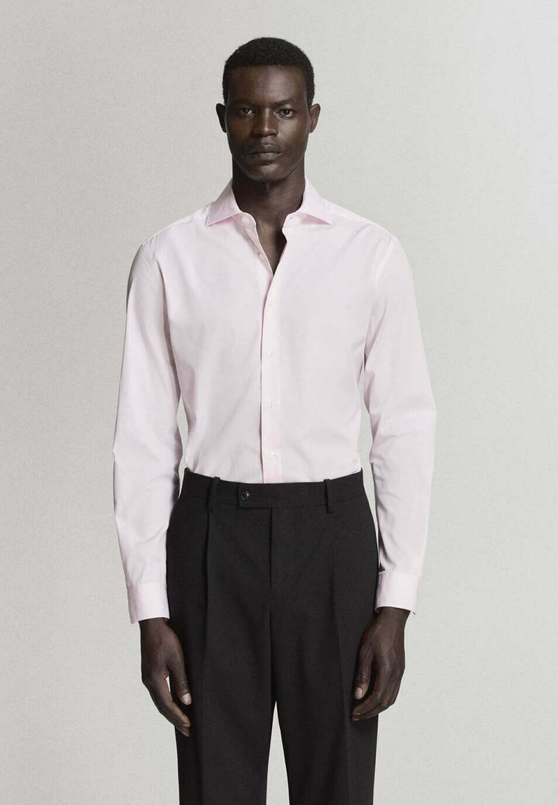 Homme portant une chemise rose pâle à manches longues rentrée dans un pantalon noir taille haute, debout devant un fond clair uni.
