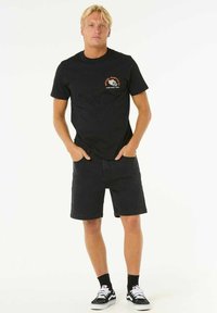 Camiseta de algodón negra con un pequeño gráfico en el frente, combinada con pantalones cortos de mezclilla negros. El modelo lleva zapatillas negras y blancas, con calcetines negros.