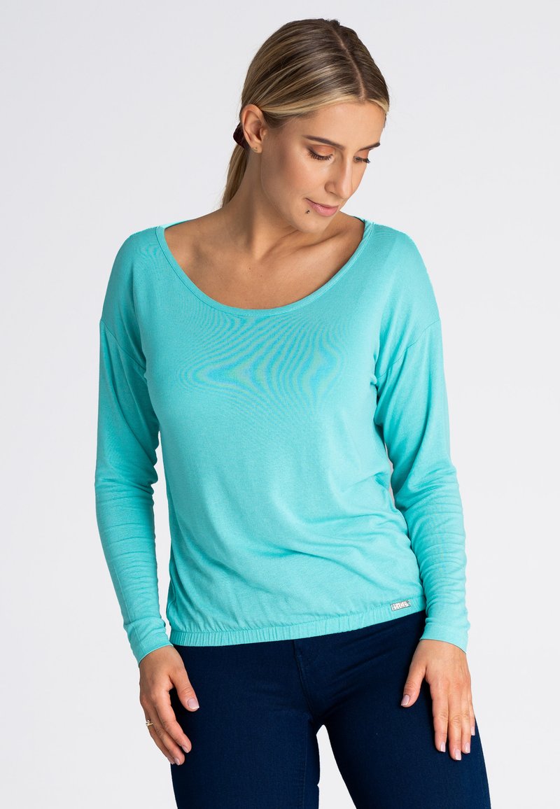 Turkusowa bluzka z długim rękawem, szerokim dekoltem i marszczonym dołem, wykonana z miękkiego materiału, zestawiona z ciemnoniebieskimi dżinsami.