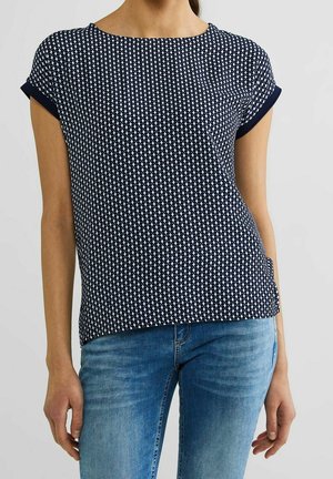 Vrouw draagt een navyblauwe top met korte mouwen en een wit geometrisch patroon en lichtblauwe spijkerbroek, staand tegen een effen lichtgrijze achtergrond.