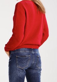 Maglione lavorato a maglia rosso con texture e maniche lunghe, abbinato a jeans in denim blu con design della tasca posteriore e finitura sbiadita.