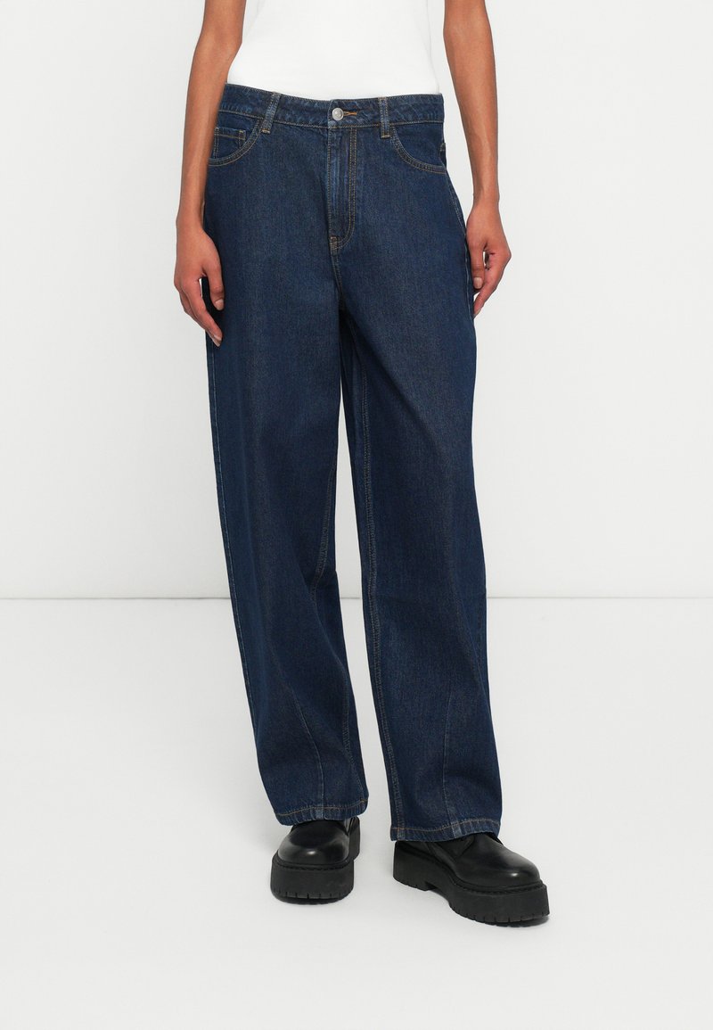 Personne portant un jean large bleu foncé, un haut blanc et des chaussures noires à semelle épaisse, se tenant contre un fond blanc uni.