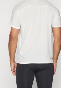 Vitt atletiskt kortärmat linne med texturerad yta, lätt mesh-design, sidoslitsar och liten Nike-logotyp på nedre delen av ryggen.