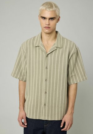Chemise à manches courtes à boutons de couleur vert olive avec des rayures verticales blanches. Elle présente un col, un tissu texturé et une fermeture à boutons sur le devant.