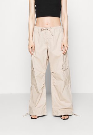 Femme portant un pantalon cargo beige à jambes larges avec taille à cordon et un crop top noir, debout pieds nus sur un fond uni.