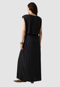 Zwarte maxi-jurk met kapmouwen en een geplooide taille. Zachte stof met een gladde textuur. Draagt met een patroon, puntige ballerina's en een gouden armband.