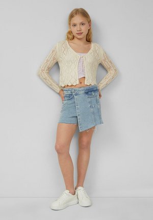 Junge Frau trägt eine cremefarbene, gestrickte, kurz geschnittene Strickjacke über einem gerafften lila Top, asymmetrischem Jeansrock und weißen Sneakers, steht an einer grauen Wand.