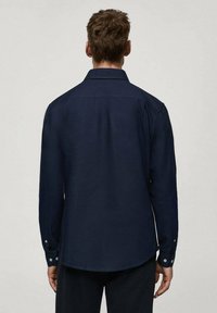 Camicia a maniche lunghe blu navy con colletto strutturato, polsini con bottoni e orlo dritto. Il tessuto appare testurizzato e resistente.