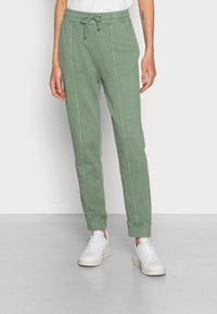 Pantalones de chándal verdes hechos de tela suave, con cintura ajustable mediante cordón, paneles laterales y puños elásticos. Llevados con zapatos deportivos blancos.