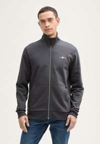 Chaqueta gris con cremallera y alto cuello, confeccionada en una tela suave. Presenta dos bolsillos frontales y un pequeño logo con el año "1962" en el pecho.