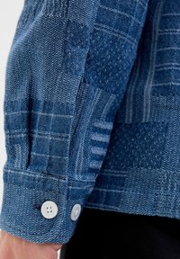 Džínová košeľa s textúrovaným modrým vzorom, ktorá má dva biele gombíky na manžete a štruktúrovaný, upravený okraj pri zápästí.