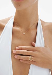 Collar de oro con un pequeño colgante ovalado, llevado sobre piel desnuda, combinado con varios anillos de oro en los dedos que tocan la clavícula. Atuendo blanco.