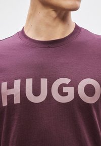 Lilla puuvillane T-särk, mille ees on suur heledat värvi "HUGO" tekst, ümmargune kaelus ja lühikesed varrukad. Sile tekstuur.