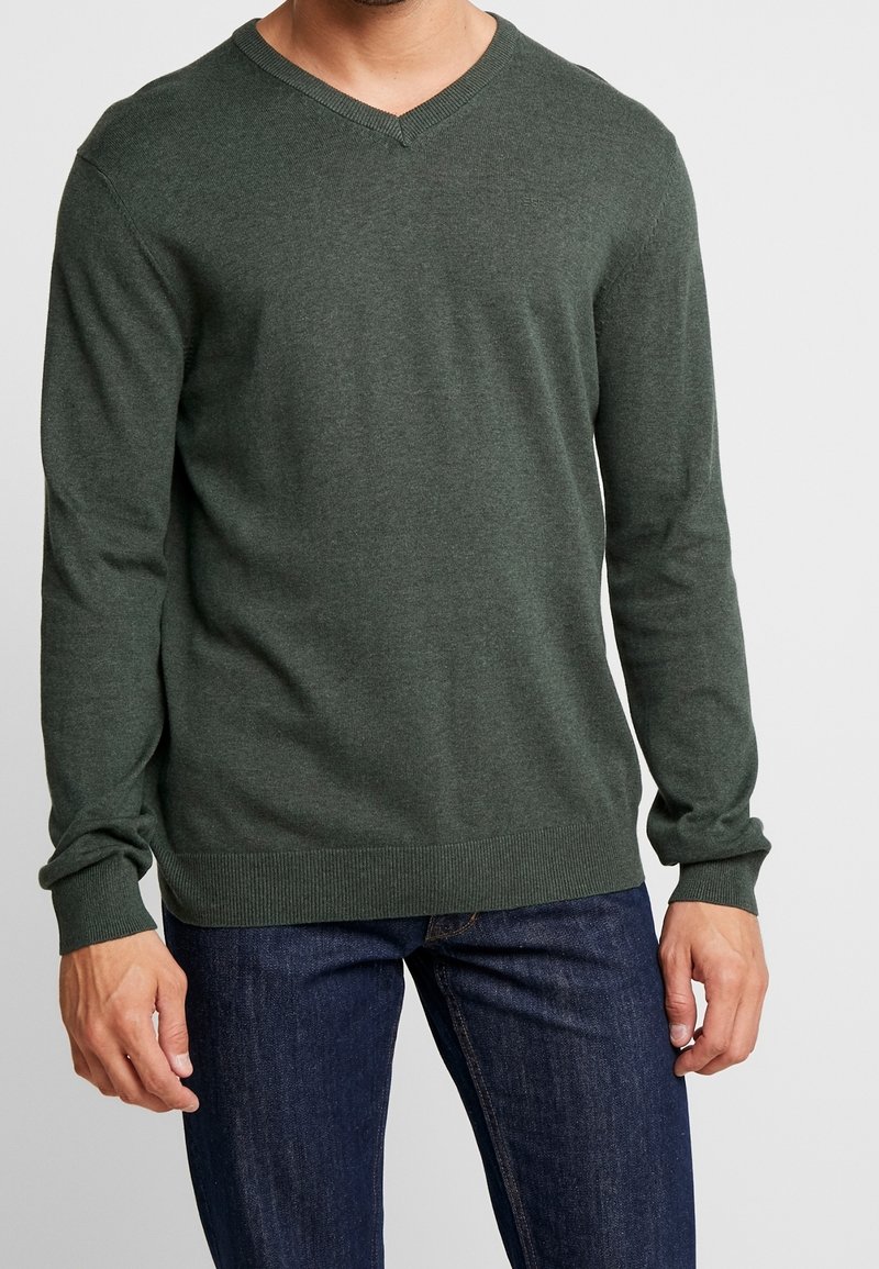 Homme portant un pull en V vert foncé à manches longues et un jean bleu foncé sur un fond clair uni.