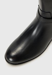 Close-up of a shiny black boot showing the toe and lower shaft, featuring a sleek design with a stitched edge and a buckle accent.