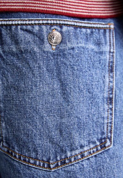 Denimstof in blauw, met een achterzak met een zilveren metalen knoopdetail. De textuur is licht gekreukt. Gestikt met oranje draad.