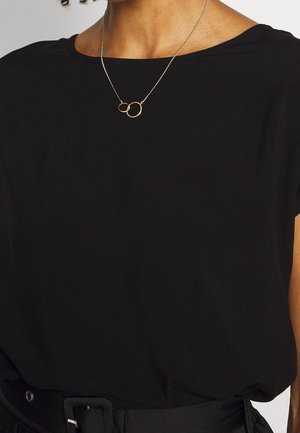 Persoon draagt een zwart topje en riem, met een gouden ketting met twee in elkaar grijpende ringen aan een dunne ketting.