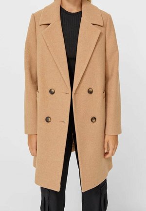 Femme portant un manteau croisé en laine camel par-dessus un pull noir côtelé et un pantalon noir.