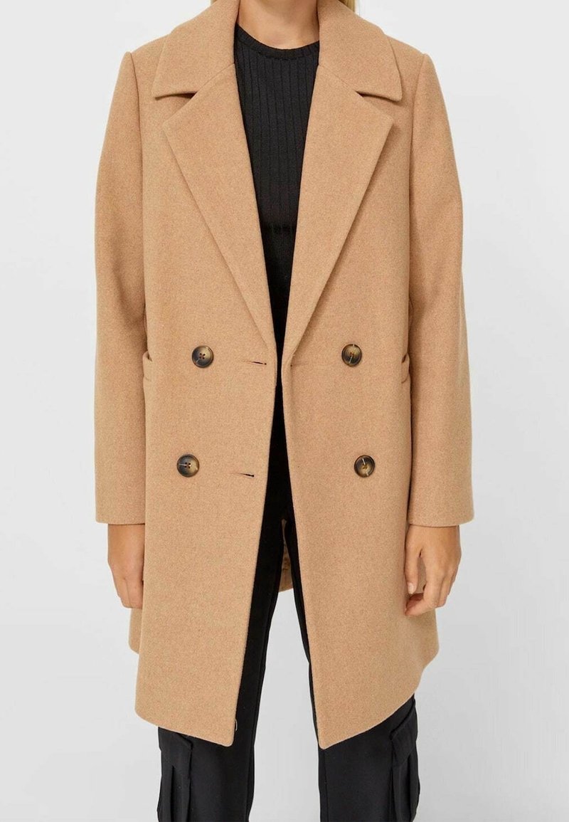 Femme portant un manteau croisé en laine camel par-dessus un pull noir côtelé et un pantalon noir.