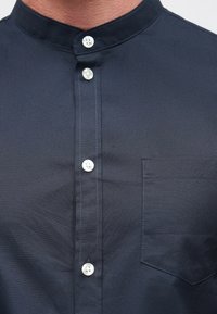 Μπλε πουκάμισο σκούρου ναυτικού με όρθιο γιακά, διαθέτει λευκά κουμπιά, τσέπη στο στήθος και λεπτή υφή �υφάσματος. Ραφές κατά μήκος της πατιλέτας.