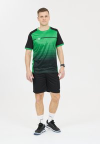 Vihreä ja musta urheilupaita, jossa on gradienttimuotoilu, yhdistetty mustiin housuihin. Päällä mustat kengät ja valkoiset sukat, joissa on logo- yksityiskohtia.