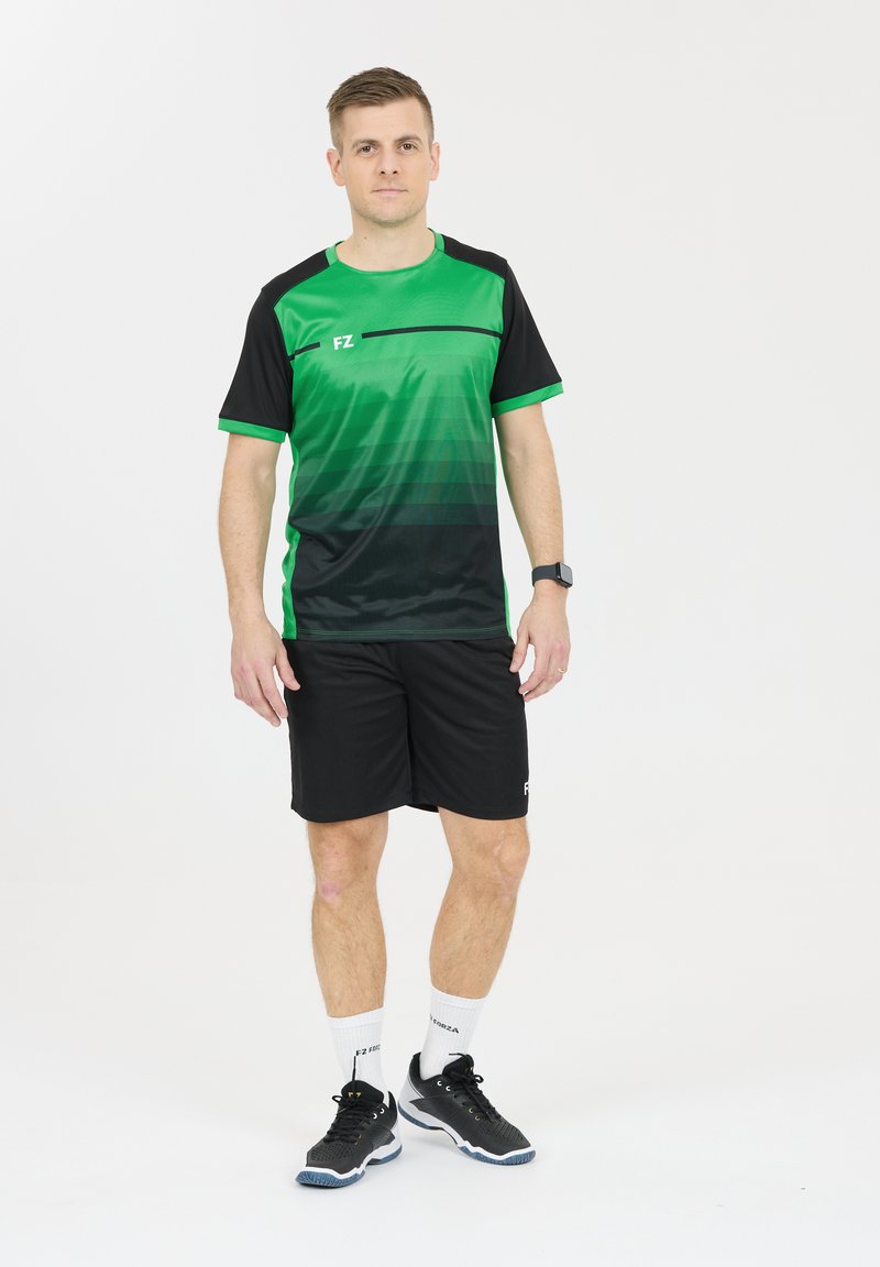 Vihreä ja musta urheilupaita, jossa on gradienttimuotoilu, yhdistetty mustiin housuihin. Päällä mustat kengät ja valkoiset sukat, joissa on logo- yksityiskohtia.