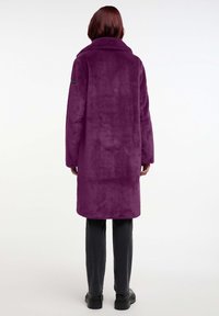 Abrigo largo de peluche púrpura con cuello, textura suave y corte relajado. Se lleva sobre pantalones negros, con un logotipo discreto en la manga.