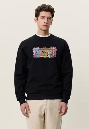 Jeune homme aux cheveux bouclés portant un sweat-shirt noir avec une scène urbaine colorée et un pantalon beige, debout devant un fond uni.