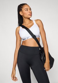 Witte sportbeha met zwart Nike-logo en verstelbare bandjes, gecombineerd met zwarte legging met hoge taille. Houdt een zwarte sportschooltas vast.