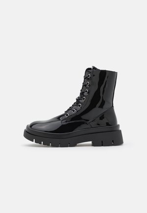 Bottes chevilles en cuir verni noir avec une semelle plateforme épaisse noire, munies de six œillets et de lacets épais. Texture lisse et brillante.