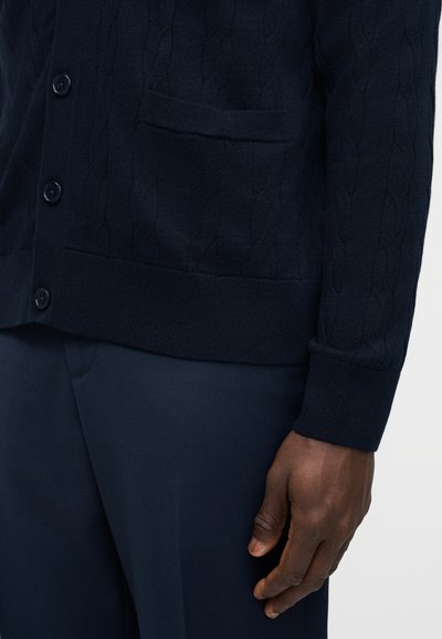 Σκούρο μπλε μπλε ζακέτα με πλέξη καλωδίου και κουμπιά πάνω από ασορτί σκούρο μπλε παντελόνι, με το δεξί χέρι ενός ατόμου να ακουμπάει δίπλα στο σώμα του.