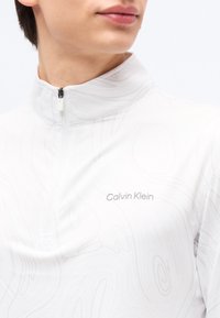 Νεαρό άτομο που φοράει ένα λευκό μπλουζάκι Calvin Klein με φερμουάρ και διακριτικά στροβιλιστικά μοτίβα, ορατό από τους ώμους έως λίγο κάτω από το πηγούνι.