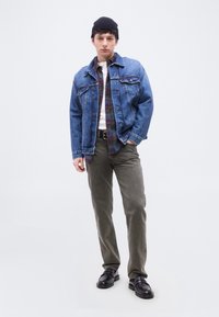 Νεαρός άνδρας με μαύρο σκουφάκι, τζιν μπουφάν με στρώσεις πάνω από καρό πουκάμισο και λευκό μπλουζάκι, γκρι παντελόνι και μαύρα δερμάτινα παπούτσια, στέκεται σε λευκό φόντο.