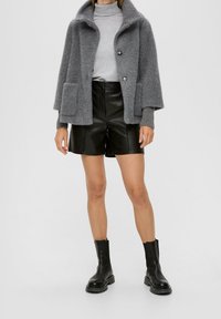 Graue Strickjacke mit übergroßem Kragen und zwei vorderen Taschen, kombiniert mit schwarzen Leder-Shorts und schwarzen Stiefeletten.