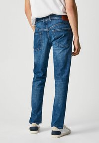 Jeans de mezclilla azules con un diseño de pierna recta, que cuentan con dos bolsillos traseros y un parche de cuero en la cintura. Llevados con zapatillas blancas.