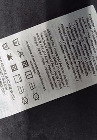 Údržbový štítok vyrobený z bielej látky, s čiernym textom a symbolmi na pranie, ktoré naznačujú pokyny na starostlivosť a zloženie materiálu.