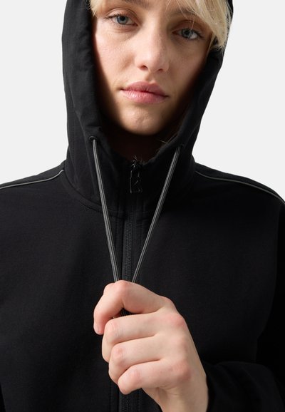 Nærbillede af person med lyst hår iført sort hættetrøje med lynlås, der holder og justerer hætte-snørebåndene.