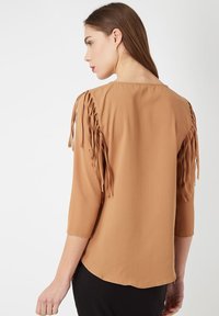 Blusa marrón con mangas tres cuartos, que presenta detalles de flecos en los hombros. Textura suave, dobladillo redondeado y diseño simple.