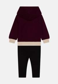 Completo con cappuccio composto da una parte superiore bordeaux, rifiniture crema e pantaloni neri. Tessuto morbido, vestibilità comoda e polsini elastici su maniche e caviglie.
