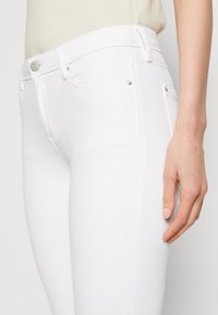 Jeans de mezclilla blanca con un corte ajustado, que cuentan con herrajes metálicos, bolsillos delanteros y una textura suave. Vista cercana de la cinturilla y la pierna.