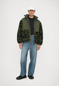 Giacca a vento verde con motivi geometrici neri, t-shirt nera, jeans di un blu chiaro e scarpe nere. Un cappello bianco completa il look.