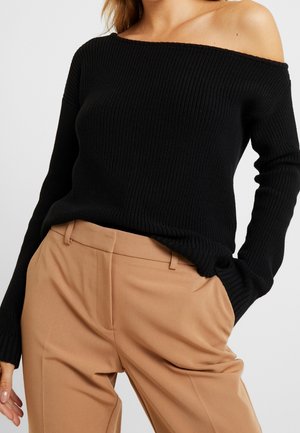 Femme portant un pull côtelé noir à épaules dénudées et un pantalon camel taille haute, une main dans la poche.